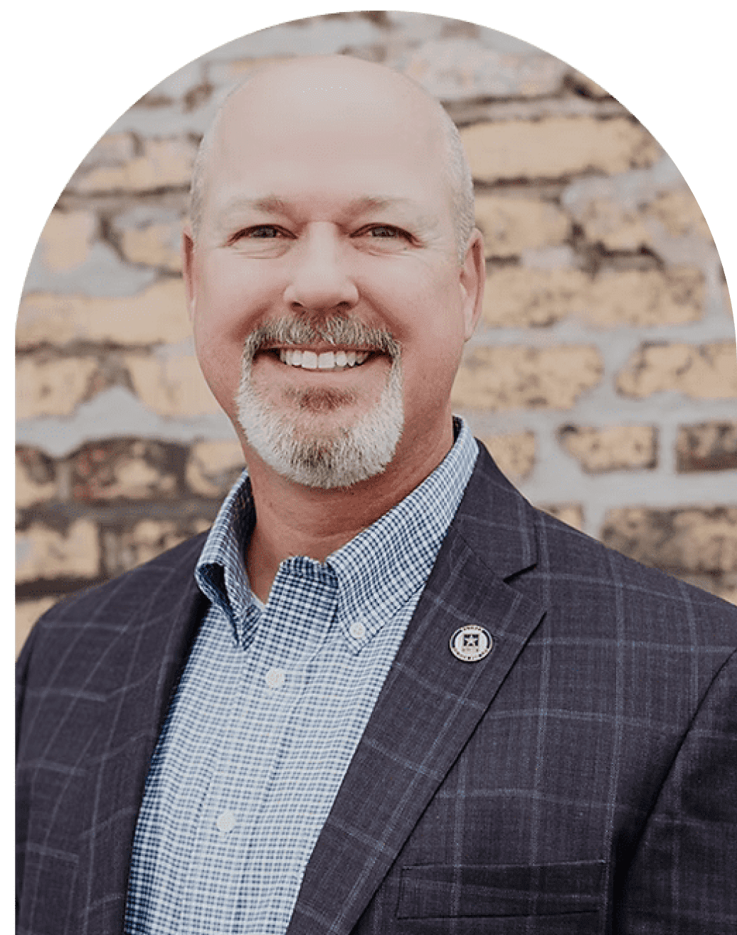 Smiling man in a checkered blazer and blue shirt against a brick wall.